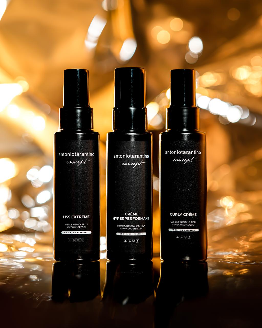 Tre flaconi neri della linea antoniotarantino concept — Liss Extreme, Créme Hyperperformant e Curly Créme — fotografati su fondo di luci dorate