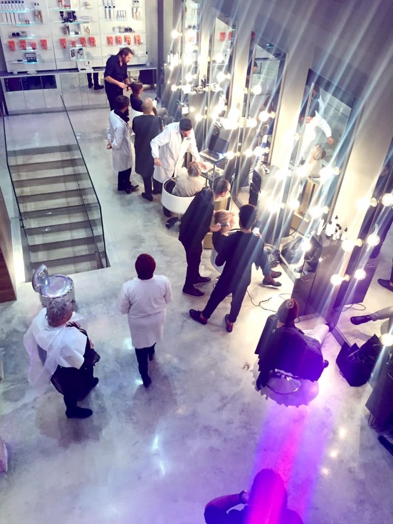 Vista dall'alto della sala del salone: postazioni con specchi a lampadine, pavimento in cemento levigato e la squadra al lavoro sui clienti