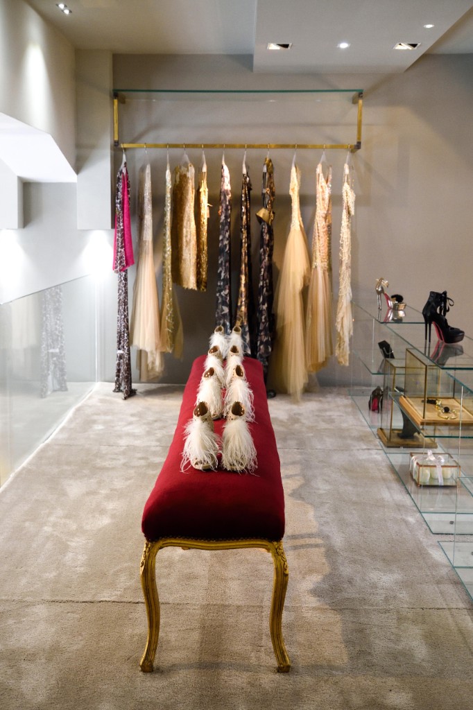 Showroom con abiti da sera appesi, panca in velluto rosso e scarpe esposte su mensole di vetro